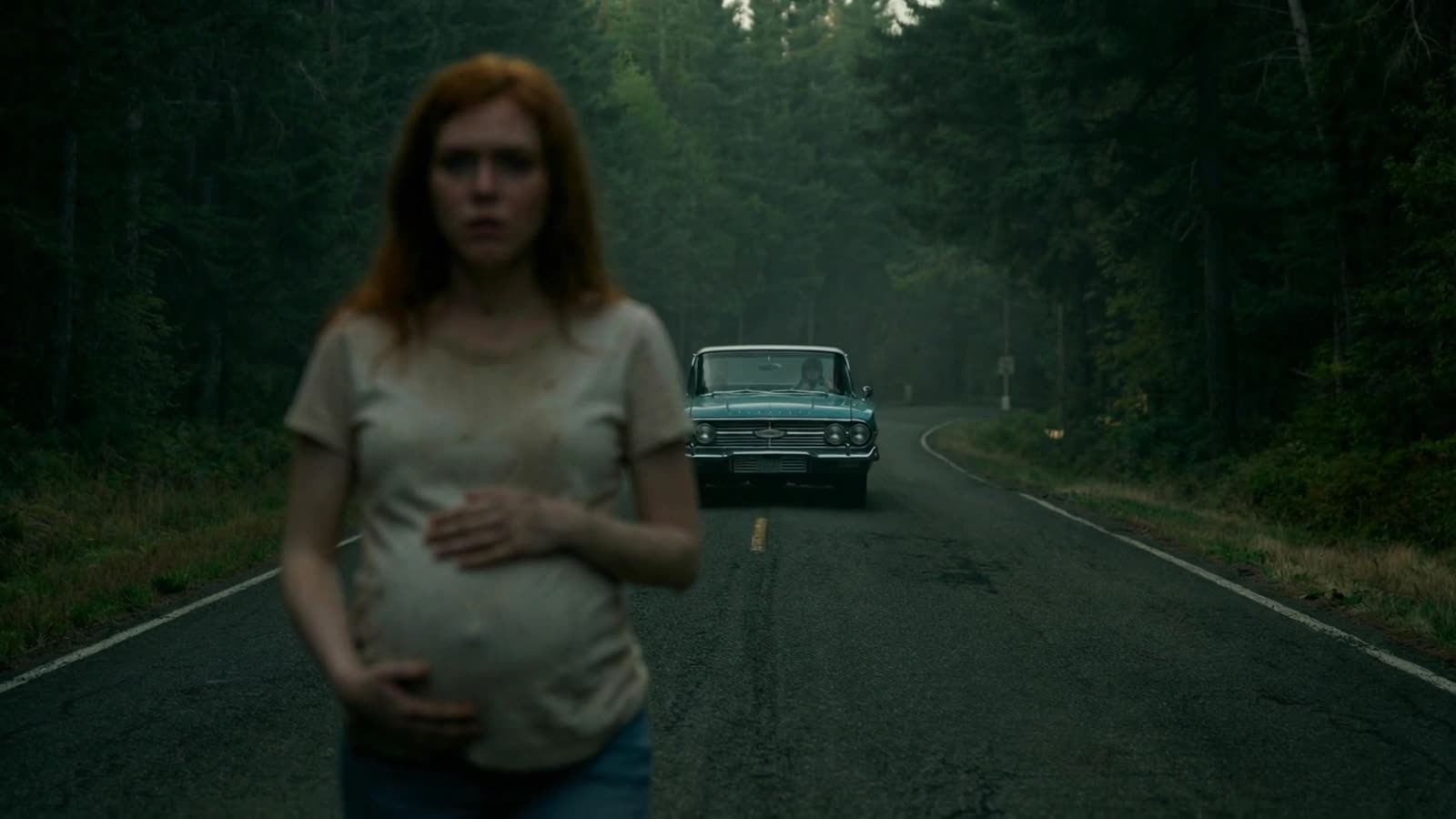 A pregnant woman stands on a forest road as an old car approaches through the mist
